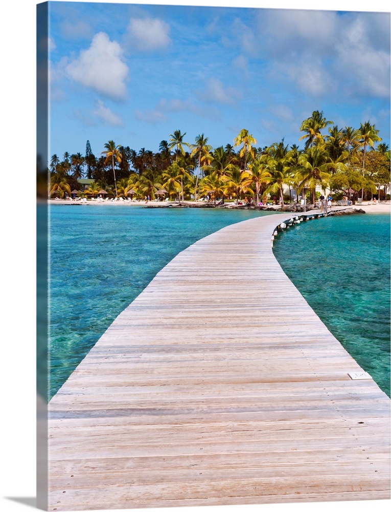 Vertical photograph on a big canvas of a wooden path over clear blue waters, leading toward a tropical island covered in p...