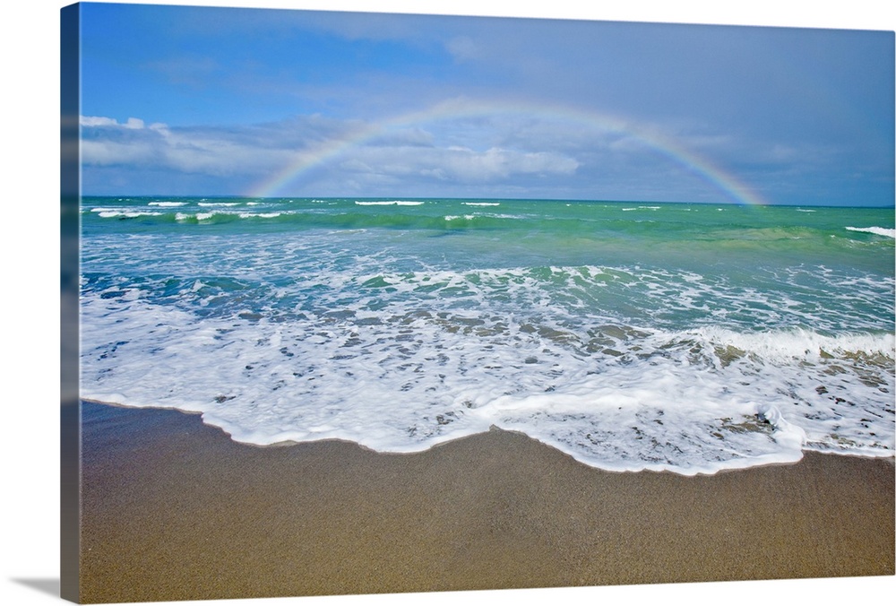 A large rainbow is photographed over the teal colored ocean.