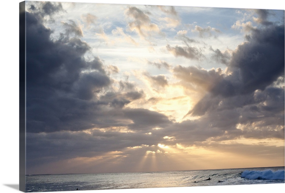 Crepuscular rays of light break through the sky over the blue ocean, surfers in the background
