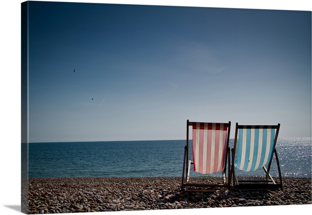 Red and blue striped deckchair and sit facing out to blue sea. Pebble beach and clear blue sky.