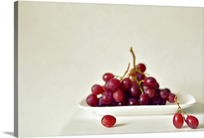 Red grapes on white square plate on white table against white wall.