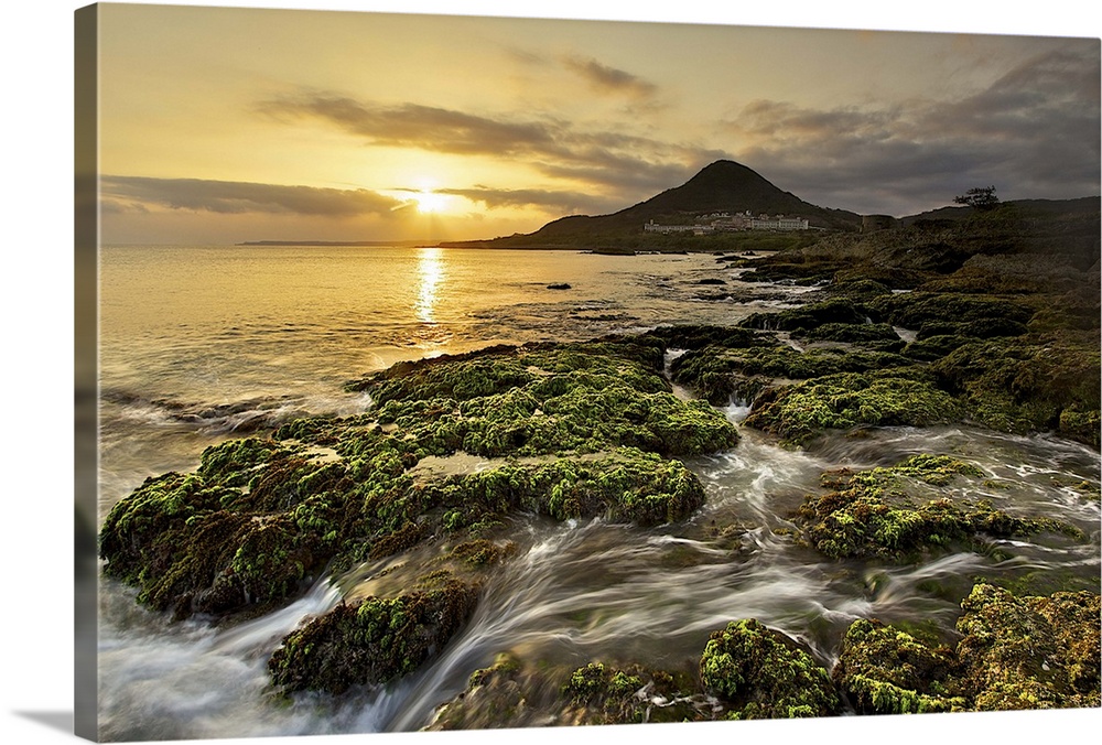 View of sailboat rock at dusk with golden sun and waves flowing down green mossy coral reefs.