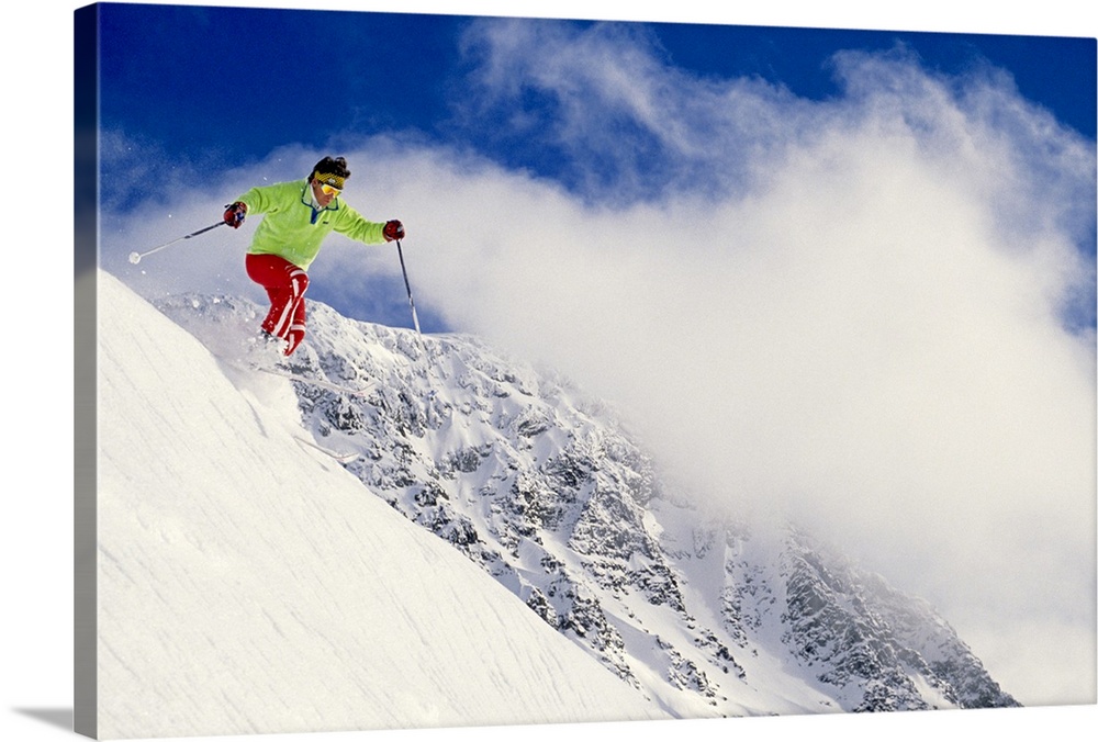 Photograph of man on skis coming down snowy slope with huge mountain in background under a cloudy sky.