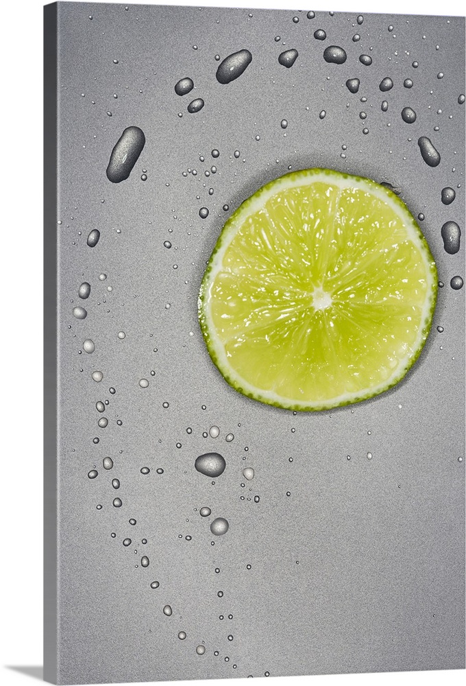 This photograph is taken of a slice of a lime with water droplets surrounding it on a grey surface.