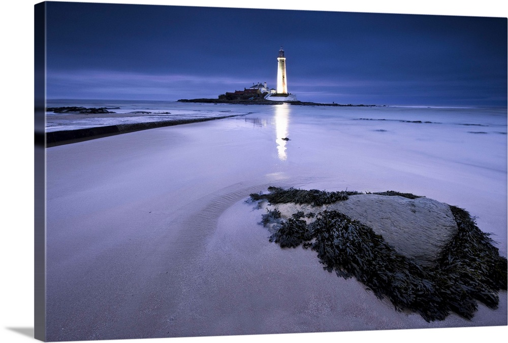 St.Marys Lighthouse, Whitley Bay, glowing at night in blue hour, reflected on sand in front of seaweed boulder.