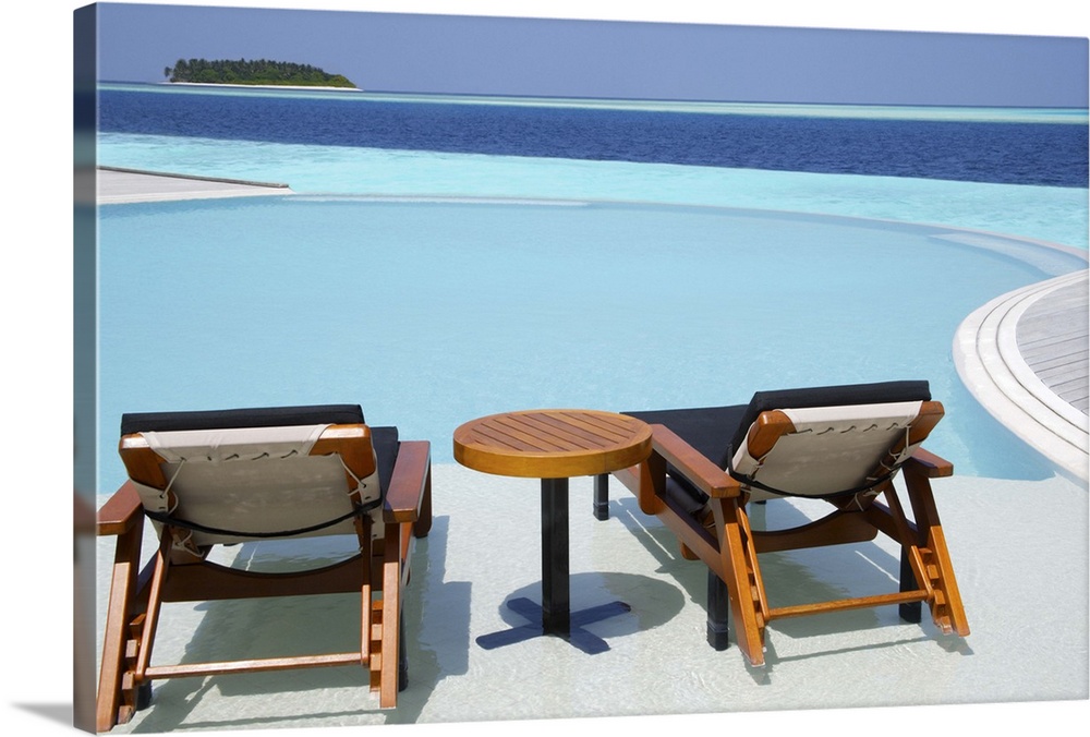 Infinity swimming pool with turquoise sea and island on the horizon. Two sun lounger chairs and table in foreground, Maldi...