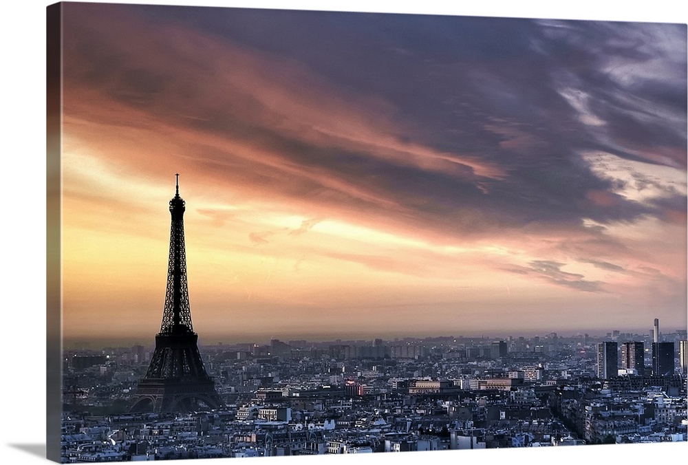 Big, horizontal photograph of the sun rising through wispy clouds as the Eiffel Tower looms over the city of Paris, France.