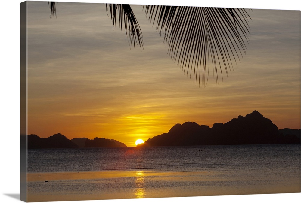 Large, landscape photograph of the sun setting behind islands, a large palm branch hangs overhead from the beaches of Coro...