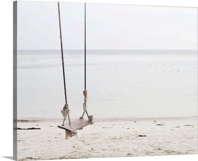 Swing made out of ropes and wood over a sandy beach directly on the ocean.