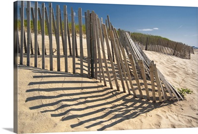 The low sun casts long shadows of a wooden fence on a sandy dune by the ocean.