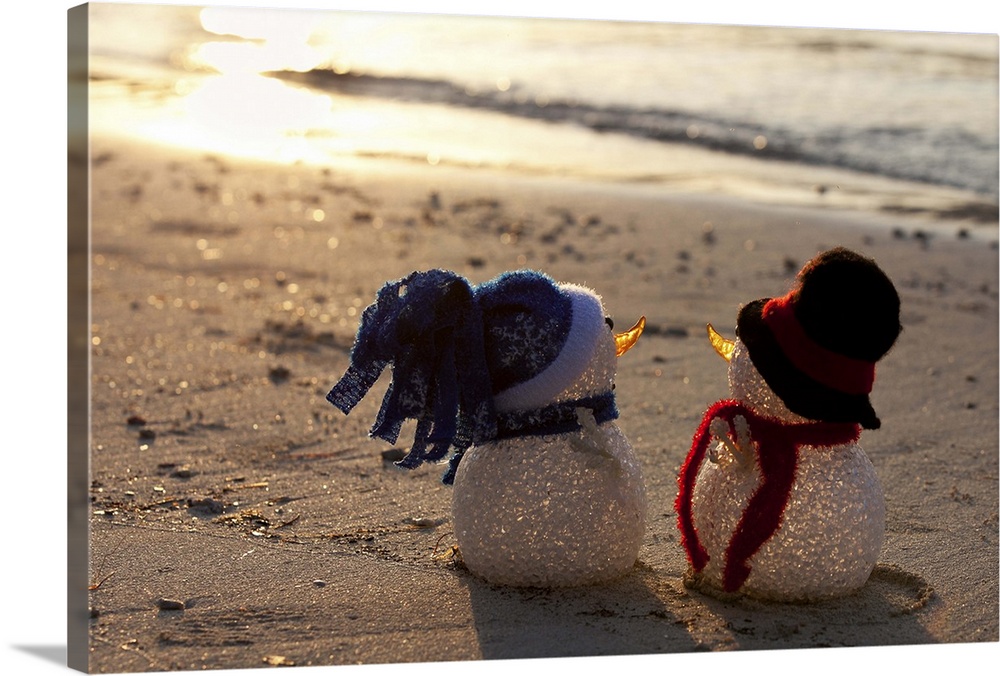 Two snowmen look to the sea at sunset. White sand and waves in the background. View from behind. Christmas decorations. Bl...