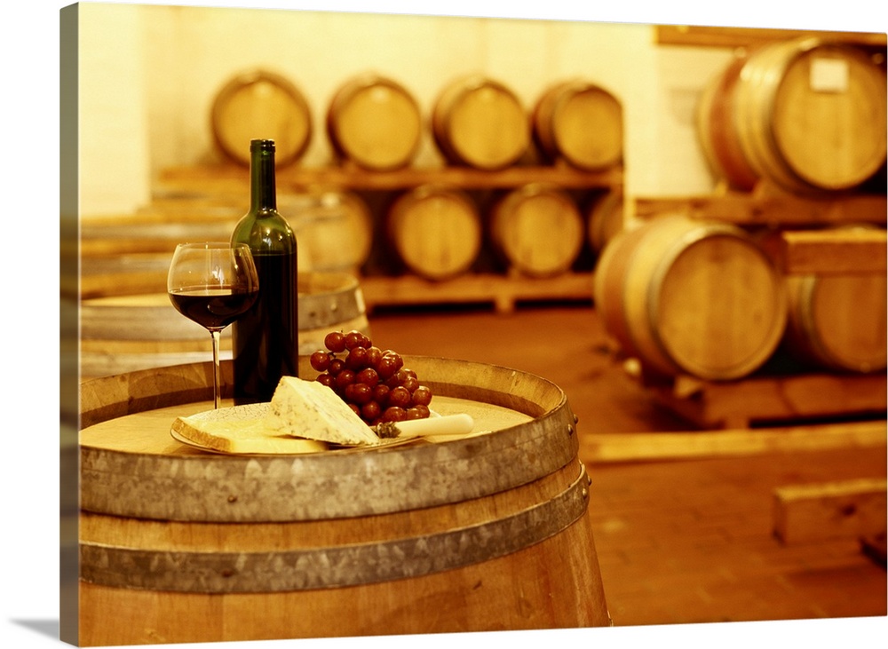 A photograph in a wine cellar of an upright barrel with a glass of wine, grapes, and cheese sitting on the table.