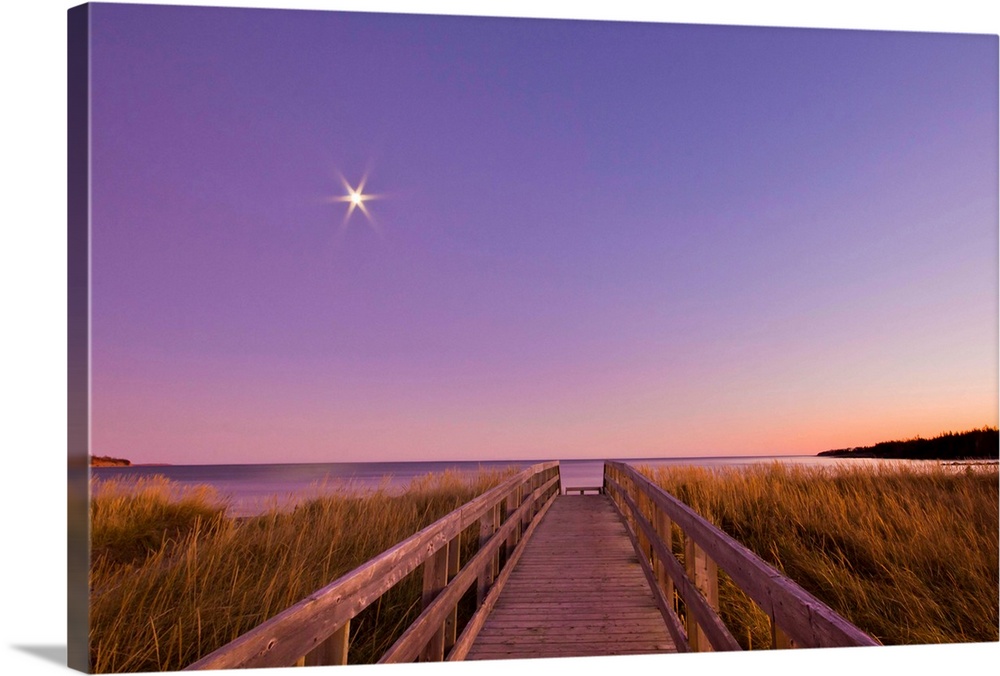 Wooden boardwalk leading through golden grasses to beach at sunset, with full moon starburst rising in clear sky.