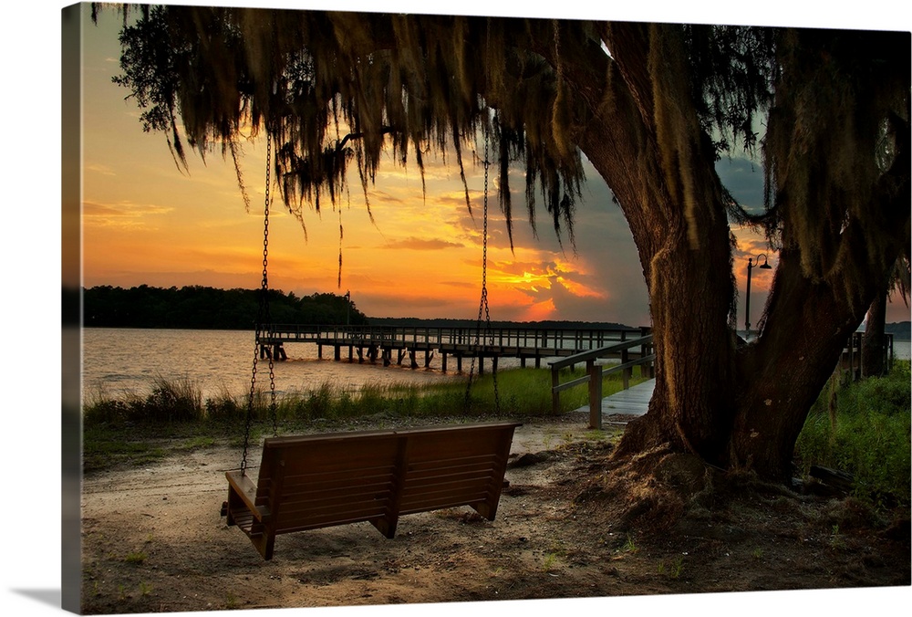 A vibrant photograph of bench swing tethered to a dropping tree, looking out over a lake at sunset.