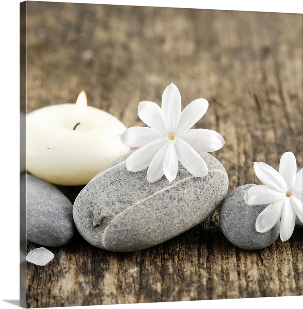 Square image of white flowers on smooth gray rocks with a candle on wood.
