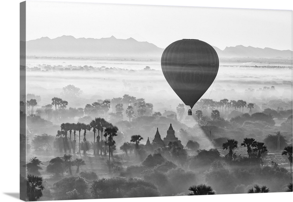A hot air balloon flies over trees and a temple at sunrise on a misty morning, Bagan, Myanmar.