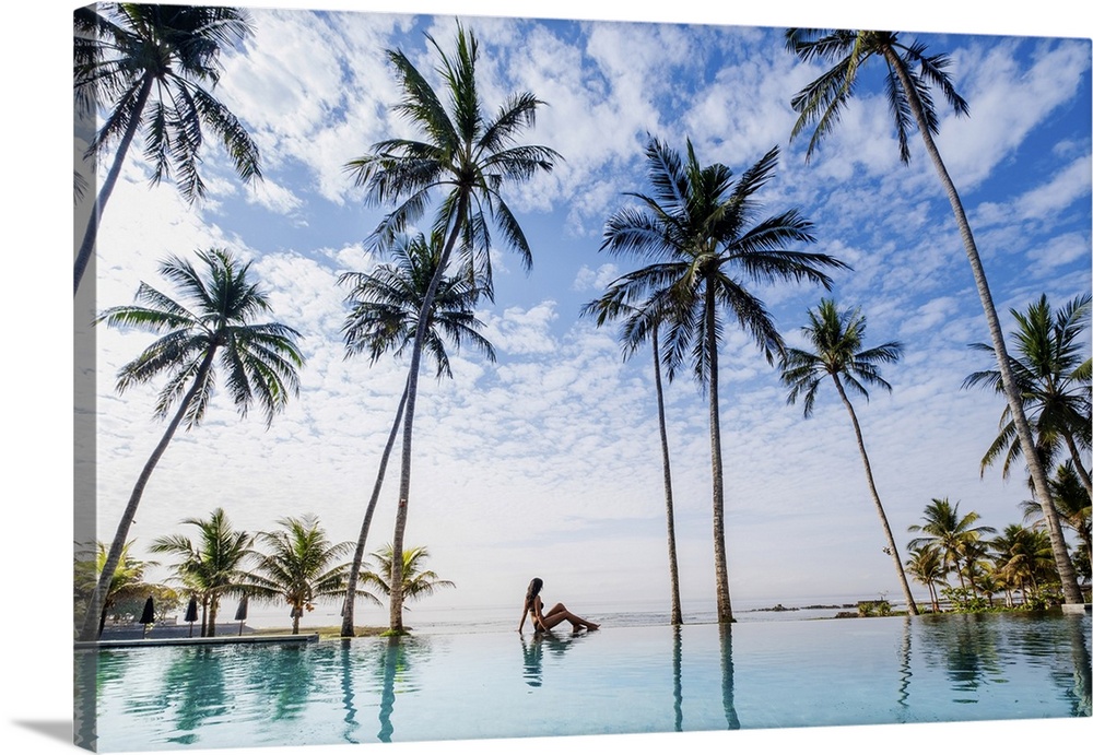 Indonesia, Bali, Candidasa. A young woman sitting on the edge of an infinity swimming pool under palm trees
