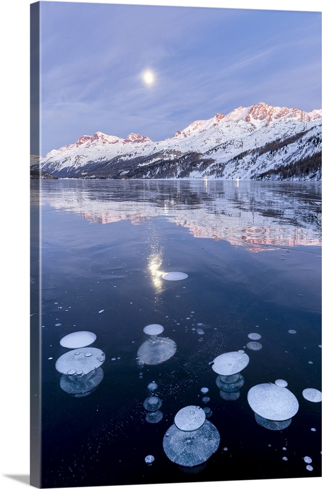 Methane bubbles in the icy surface of Silsersee with snowy peak illuminated by sunset and moon in the sky. Lake Sils, Enga...