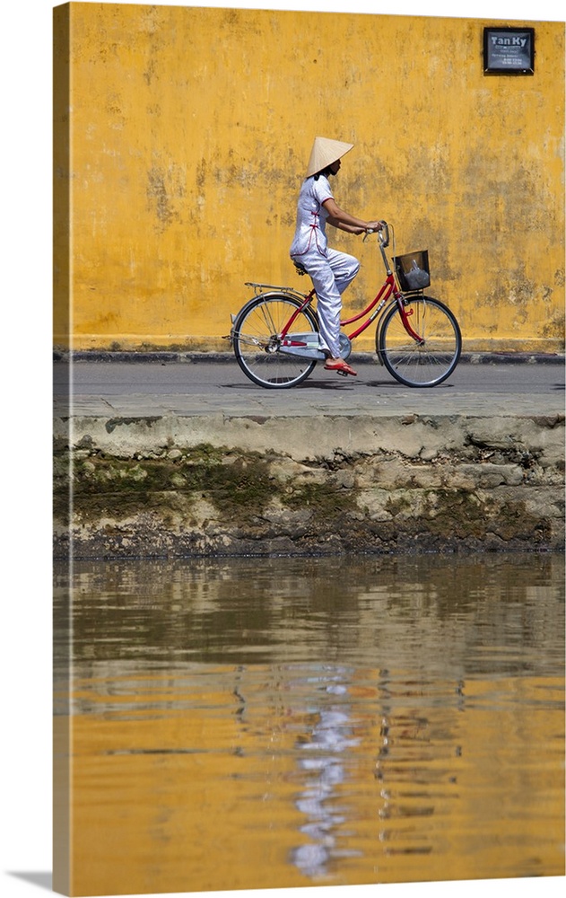 Vietnam, Quang Nam Province, Hoi An. A lady in traditional dress cycles along the waterfront of Hoi An riverfront.