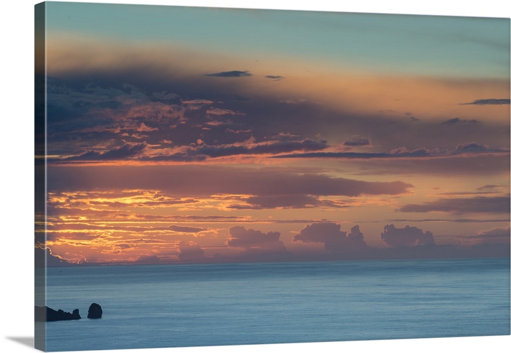 Photograph of clouds and ocean boulders casting silhouettes from the sun setting in the distance.