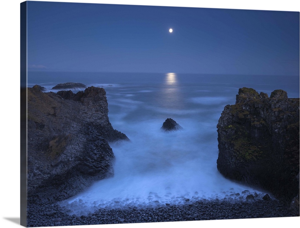 Illuminated moon in the distance hanging over the ocean in the distance, with two bookend boulders in the foreground.