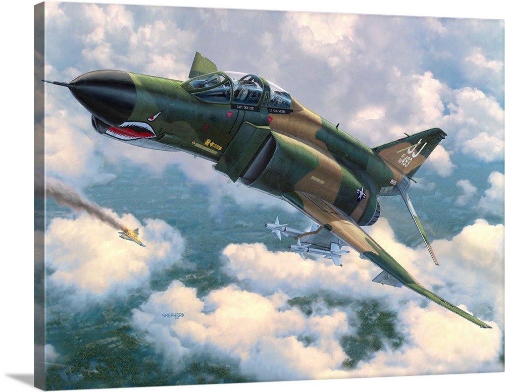 An F-4E Phantom II flies in front of broken clouds over a tree covered jungle terrain with a North Vietnamese MiG 21 going...