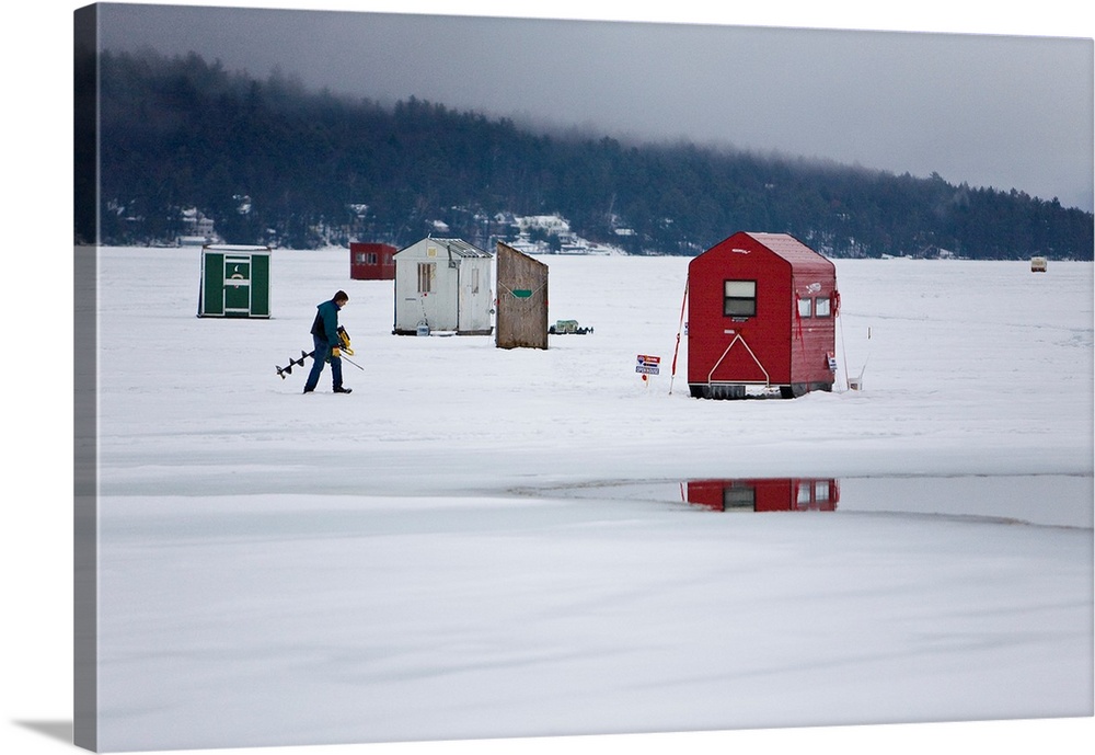 Ice fisherman with a drill walking among fishing shacks on a frozen lake.