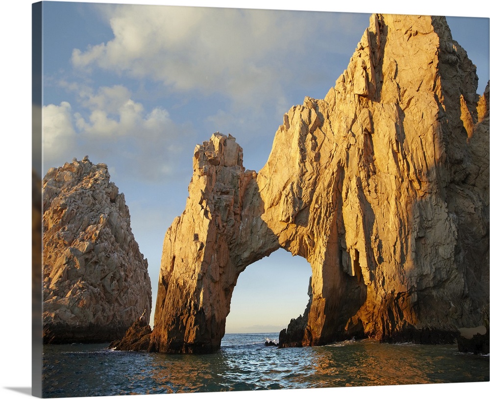 Photograph of huge rock formation with an arch in the ocean under a cloudy sky.