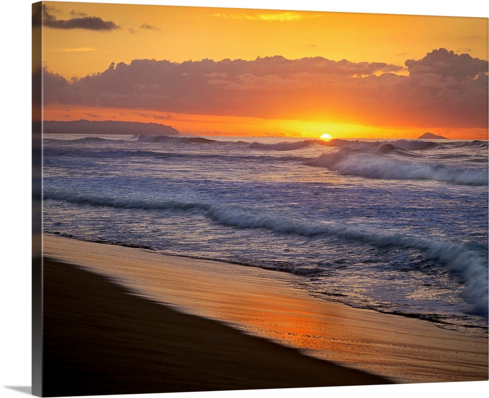 The sunset dips below the ocean horizon as ocean swells dash against the sandy tropical shoreline.