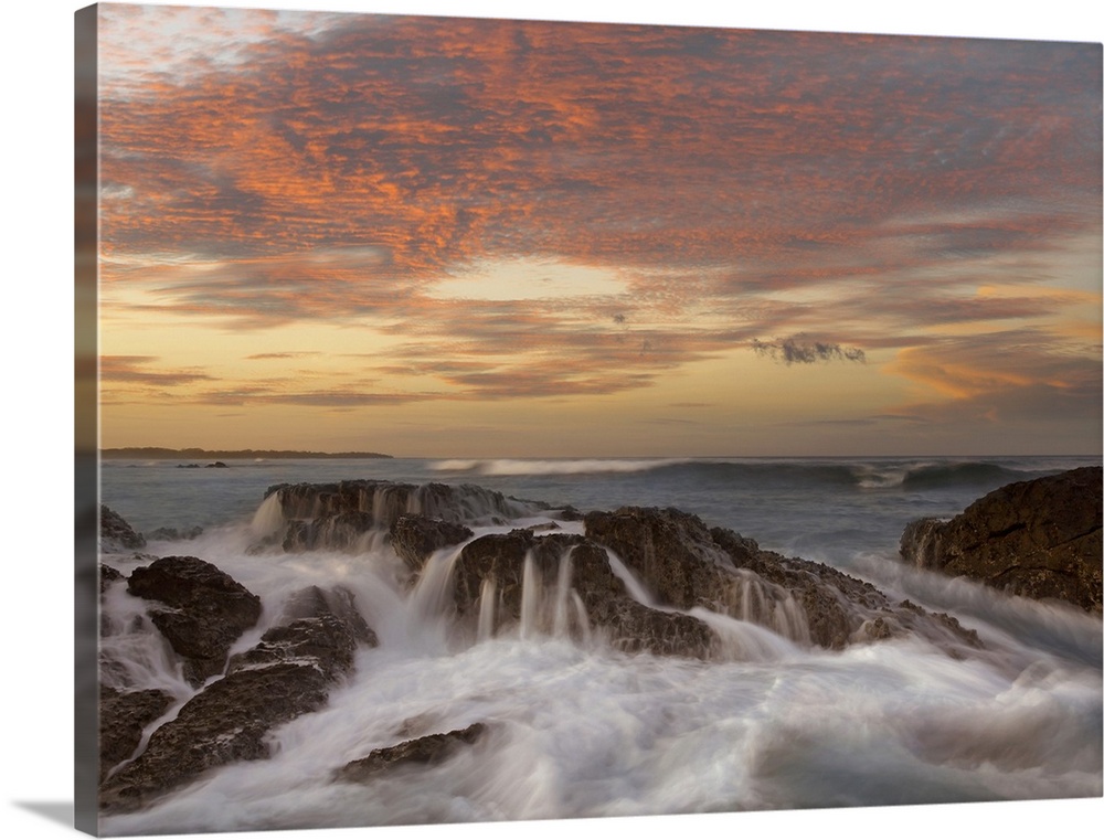 Ocean water splashing over rocks protruding from the shoreline, under a dramatic sky of clouds lit up by the setting sun.