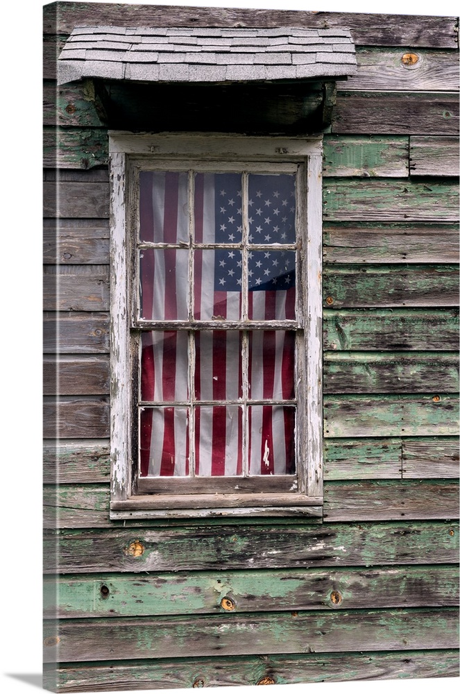 An American Flag hanging in the window of a house with weathered wooden siding.