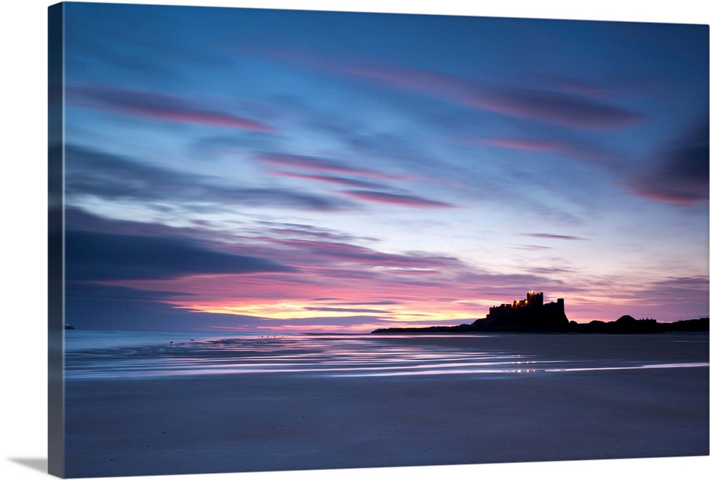 A dramatic dawn of blues, purples, oranges, reds and golden yellows over wet sand at Bamburgh Castle, Northumberland, UK.
