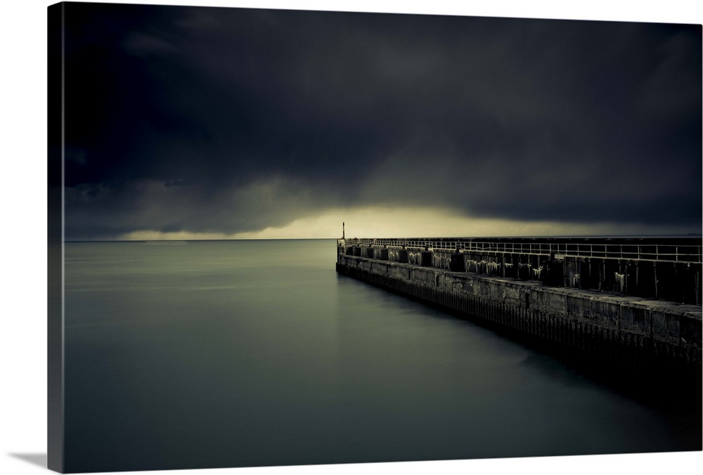 A photograph of a pier jetting out over water under a sky filled with a blanket of dark clouds.