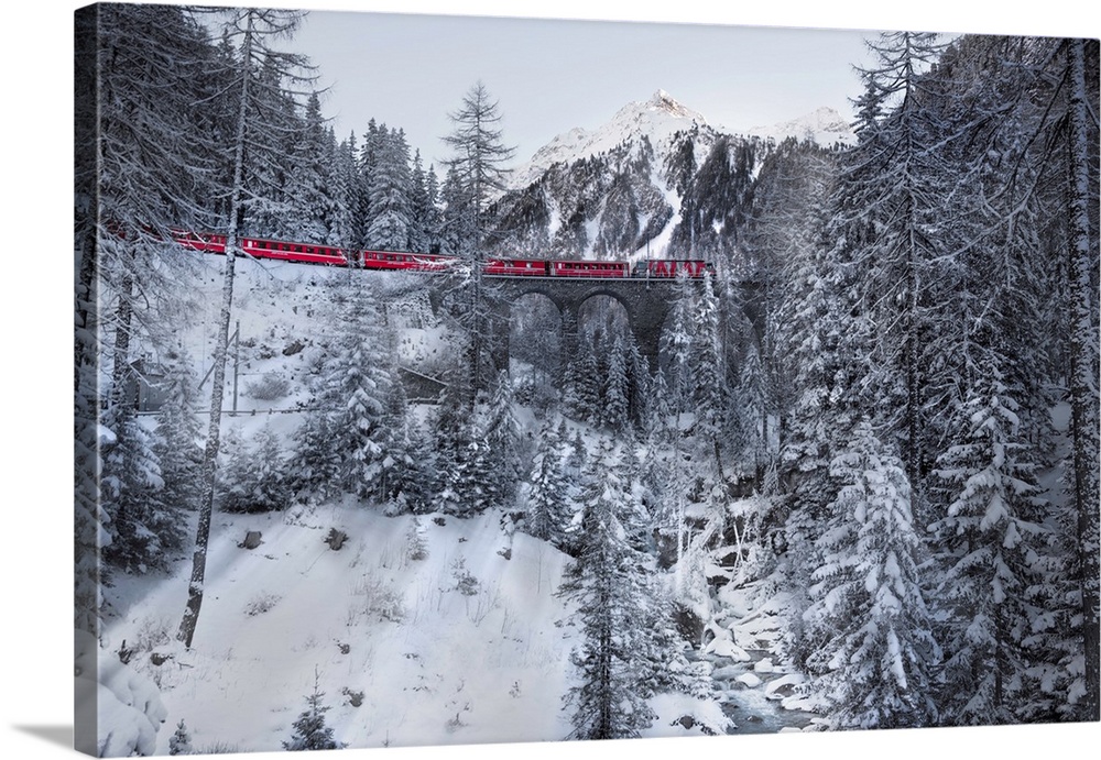 Photograph of a red train passing through a snowy mountainous valley in winter along an arched railroad.