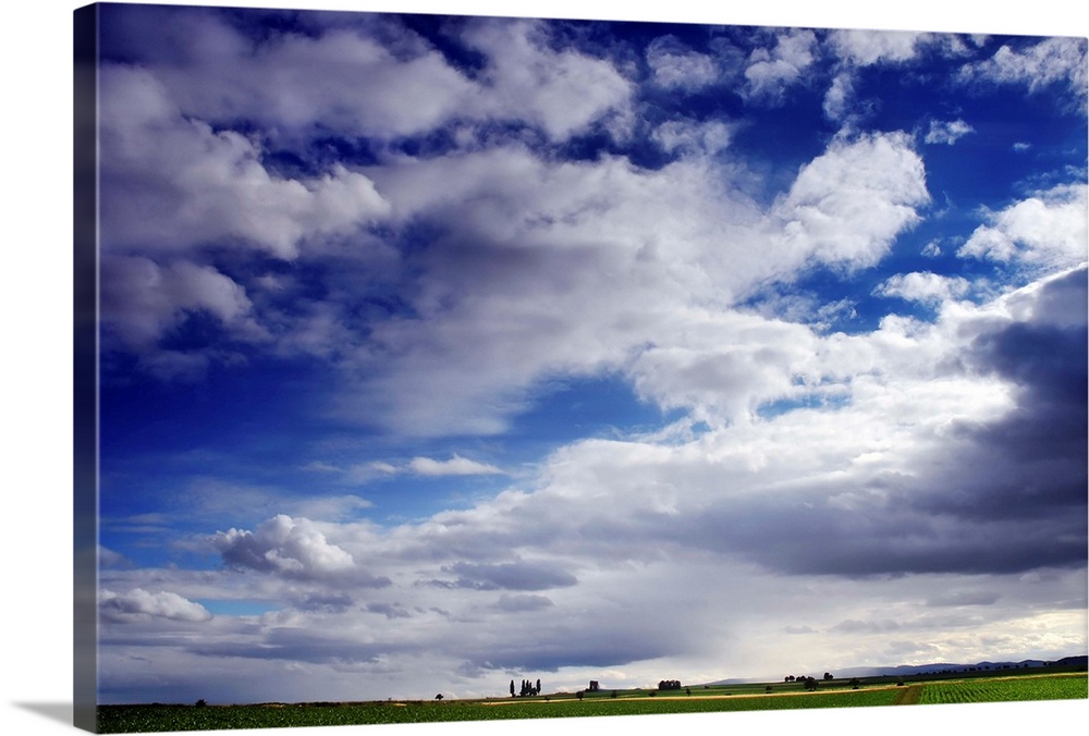Large horizontal photograph of many fluffy white clouds in a vibrant blue sky, over a green farm landscape.