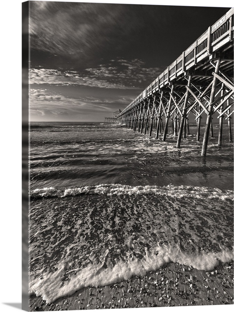 Black and white image of a pier leading into the ocean on a cloudy day.