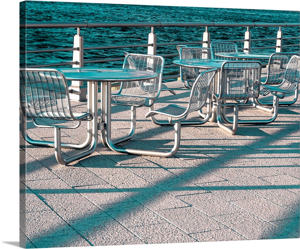 Long shadows creating abstract patterns on outdoor chairs and tables by the sea.