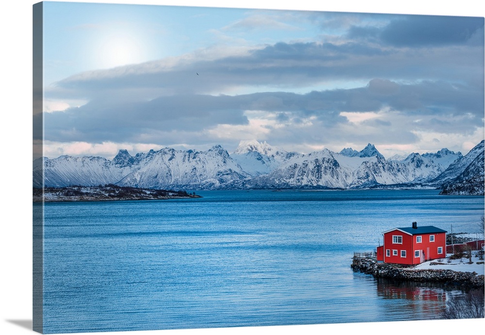 A photograph of a red building sitting on the shoreline of a snowy landscape with mountains in the distance.