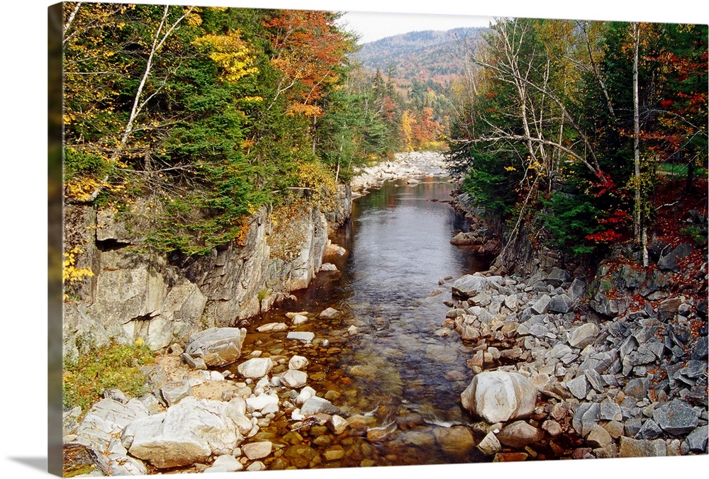 A calm creek runs through rocky terrain with forests on either side and autumn colored mountains in the background.