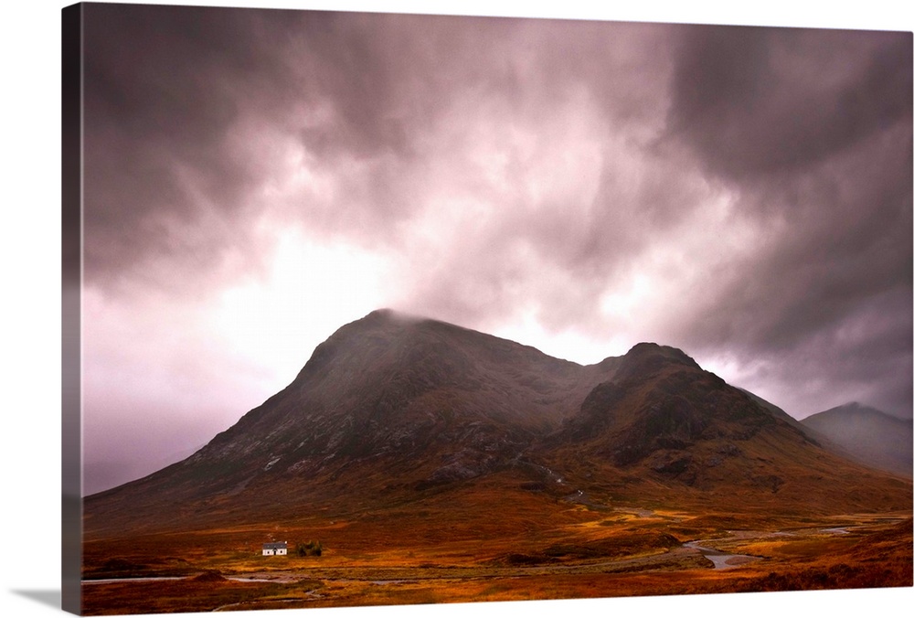 A majestic Scottish mountain in Glencoe with dramatic storm clouds and rich autumn colours with a small white cottage dwar...