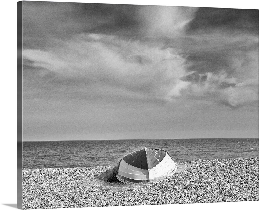 A monochrome black and white image of an upturned rowing boat on an English shingle beach in bright sunshine with fluffy w...