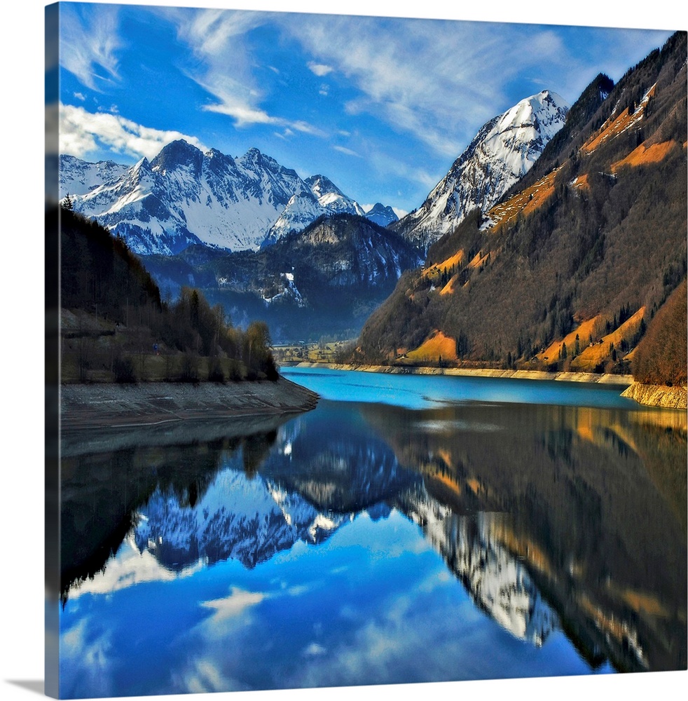 Square photograph on a big wall hanging of snow covered mountains beneath a blue sky, reflecting in still waters in the fo...
