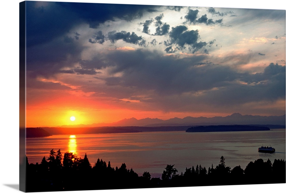 Horizontal photograph of a vibrant setting sun in a partly cloudy sky, over a large body of water, silhouetted tree tops i...