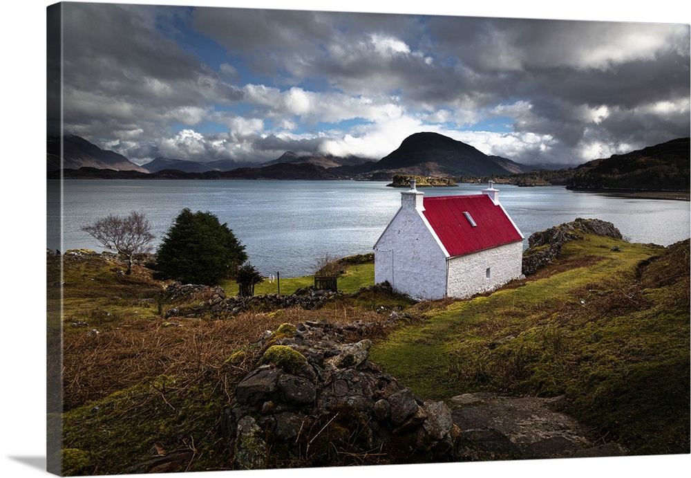 Red roofed cottage in romantic Scottish Highlands wiht blue lake and fluffy clouds in the light blue sky.