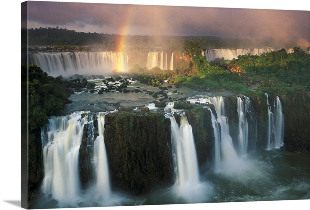 Big, horizontal, wide angle photograph of Iguazu Falls with a rainbow descending into the waters.