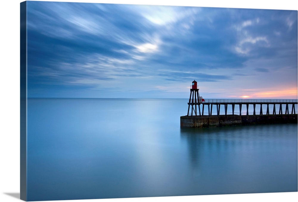 Timeless blue dawn over flat sea with scudding clouds and a lighthouse jetty.