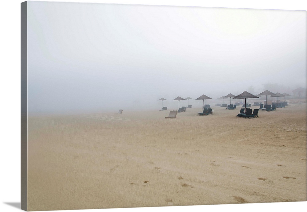 Landscape photograph of a sandy beach covered in fog and beach chairs with umbrellas.