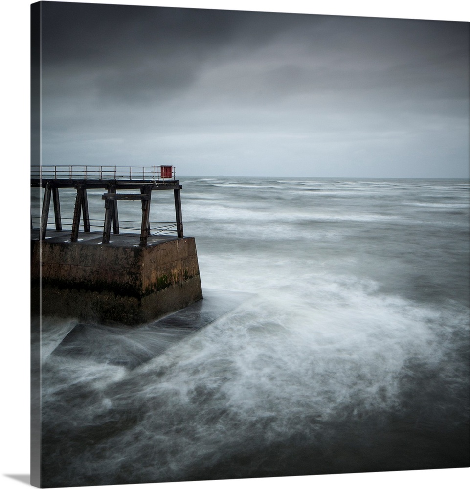 A melancholy cool blue grey image of stormy sea around a harbour installation with swirling waves and a stormy sky.