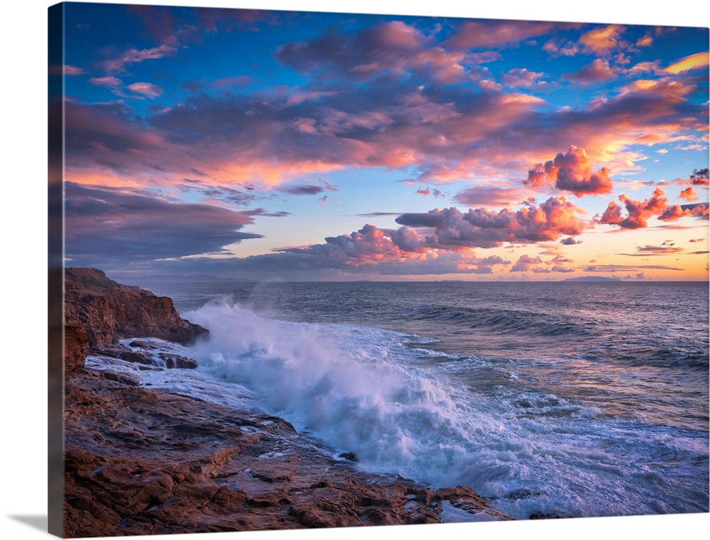 Beautiful photograph of a warm, cloudy sunset over the ocean with crashing waves onto the rocky shore.