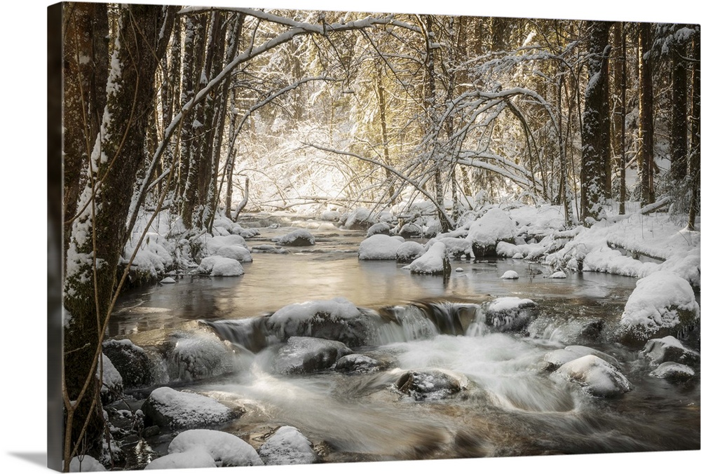 Photograph of a flowing river in the middle of the woods surrounded by snow covered rocks and trees.